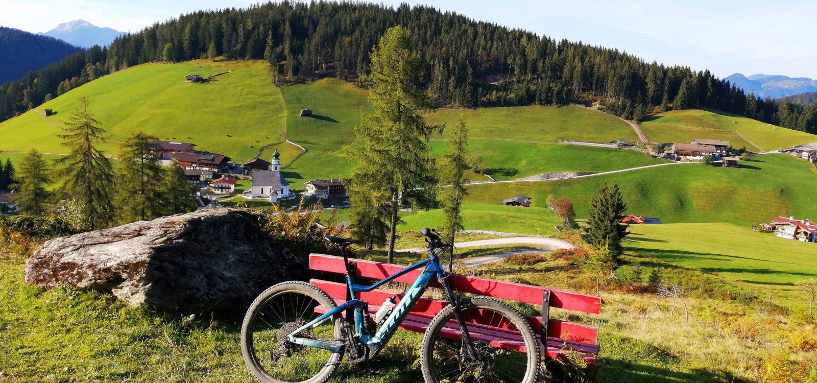 E-Bike vor roter Bank mit Blick auf Dorf, Kirche und grüne Wiesen in der Wildschönau